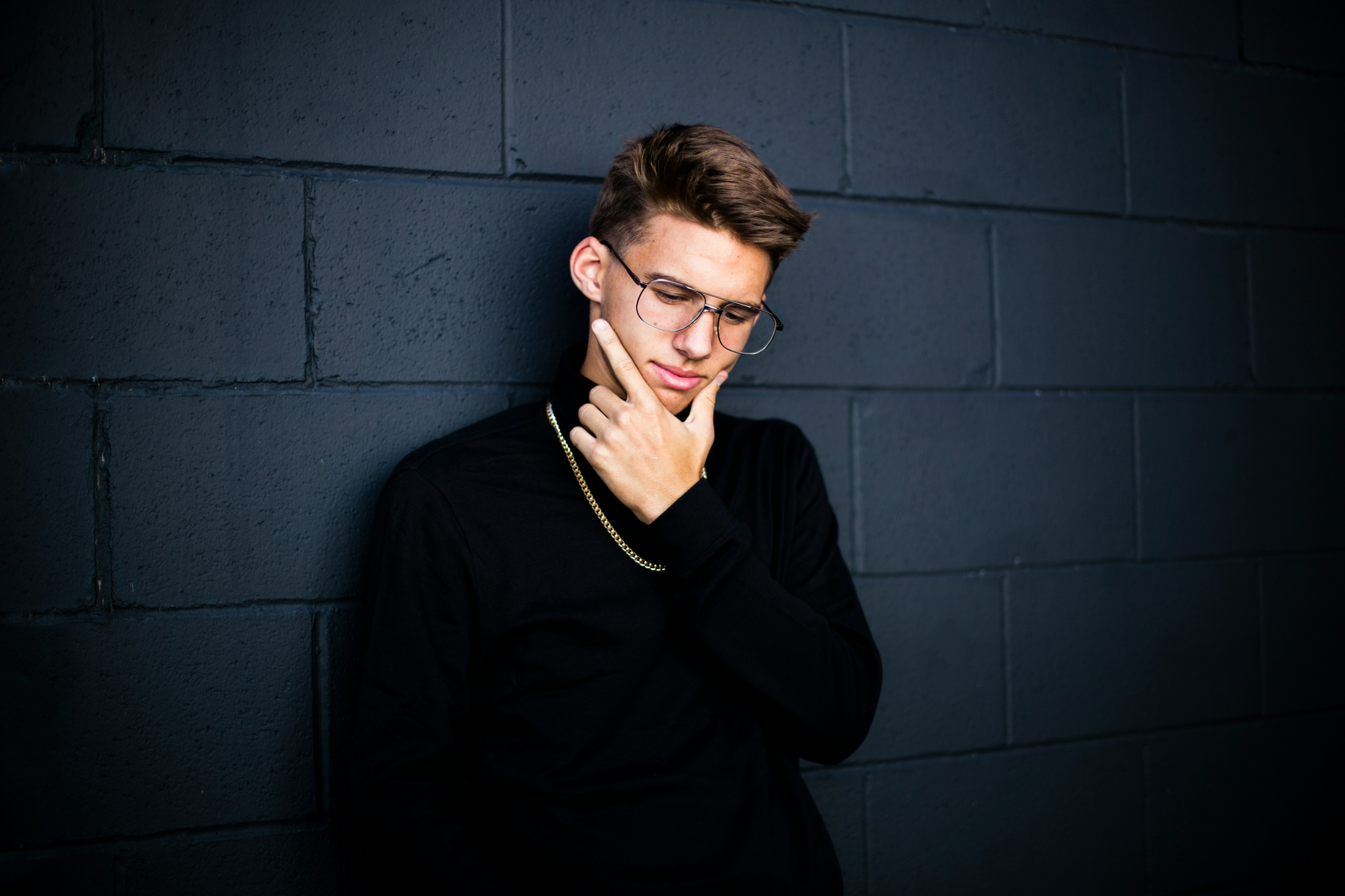 man leaning on wall while looking down wearing eyeglasses and necklace with right hand on chin